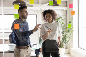 Diverse young multiethnic employees work with sticker notes develop business startup ideas on glass wall in office. Multiracial colleagues engaged in creative thinking at casual meeting. Teamwork