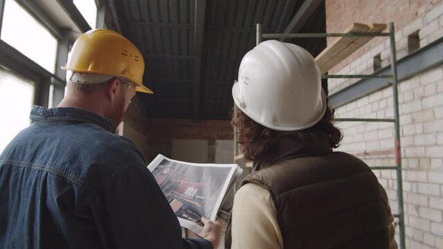 Back view of male and female engineers in hardhats standing in unfinished apartment and discussing paper slides of construction project