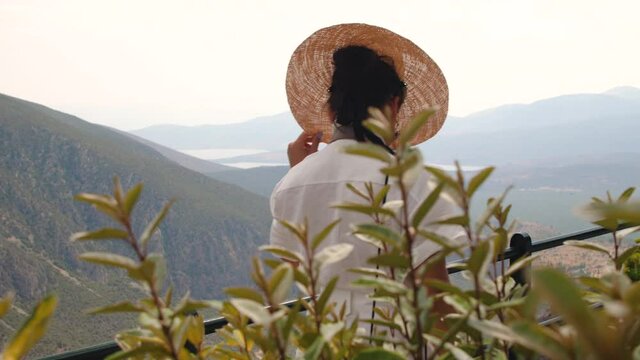 Young woman looking at Delphi landscape with mountains from high viewpoint at sunset, Greece. Fashion white dress, sunglasses, large hat.