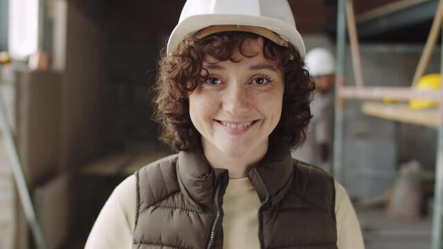 Chest up portrait shot of young female architect in hardhat standing at construction site, looking at camera and happily smiling