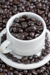 Top view of white cup and saucer with coffee beans, over more beans, selective focus, vertical
