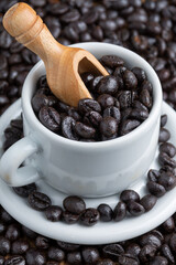 Top view of white cup and plate with coffee beans and wooden spoon, over more beans, selective focus, vertical