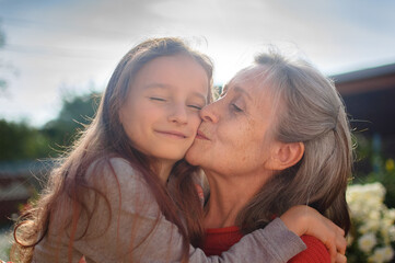 Senior grandmother with gray hair wearing red sweater with her little granddaughter are hugging in the garden and during sunny day outdoors, mother's day