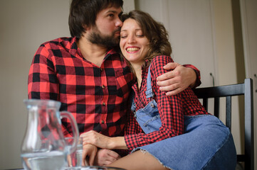 Lovely couple wearing similar red checkered shirts is hugging sitting at the table at home during celebration St. Valentine Day. Indoor portrait of boyfriend and girlfriend