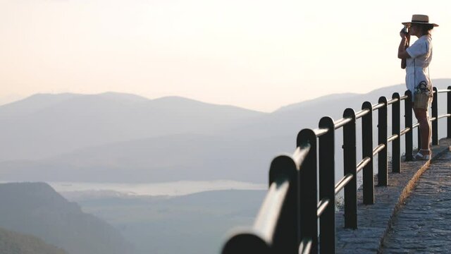 Young woman taking pictures at Delphi landscape with mountains from high viewpoint at sunset, Greece. Fashion white dress, sunglasses, large hat.