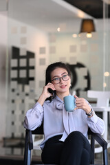 Young femal in eyeglasse talking on phone and holding a cup of hot beverage while sitting in offcie room.