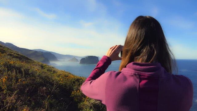 mujer joven sacando una fotografía en un lugar con vistas al mar en la zona de Bermeo mirando hacia san juan de gaztelugatxe  con bruma marina y mar calmada