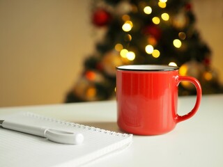 Composition of red mug with coffee, notepad and pen on the white table on the background is a Christmas tree and Christmas lights. Goals 2021. Wish list. Space for text