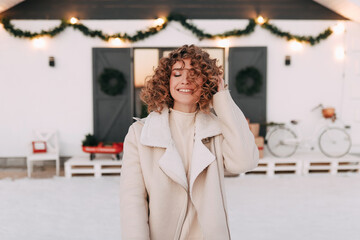 Happy cheerful beautiful cute curly haired girl with a snow white smile having fun enjoying in warm clothes looking at the camera at a decorated country house in winter