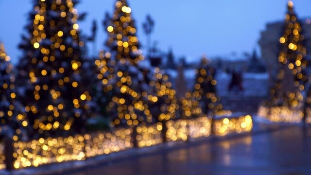 Bokeh background of blurred decorated Christmas trees in golden garland on street of evening city. Christmas and New Year holidays with fir-trees at dusk in defocus.
