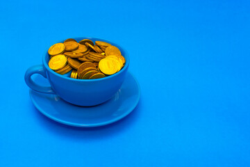 Cheapskate. Blue mug full of shining golden coins on blue saucer against blue background with blank space for text. Love of money concept. Closeup. Copy space