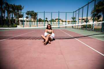 Stylish cute teenage model dressed in summer hipster vintage white stylish clothes. Trendy girl posing in sunglasses lying on a tennis court. The original shadow from the grid at the sports field