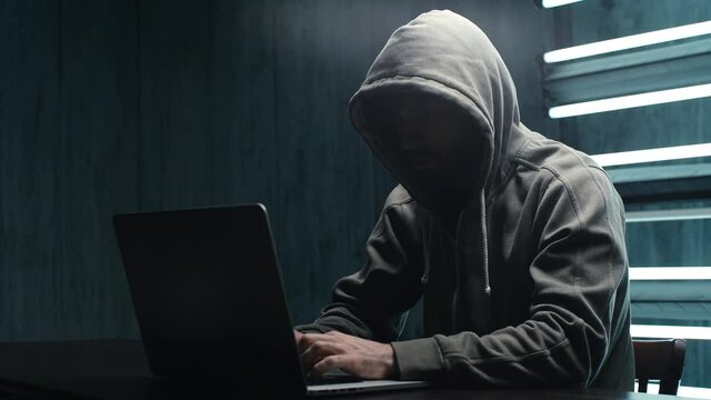 Unrecognizable man hacker wearing sweatshirt with hood typing on laptop computer keyboard and breaking password. Side view of dangerous hooded computer hacker infecting system with cyber virus.
