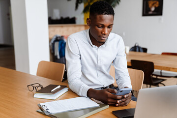 Fototapeta premium Afro american focused man using mobile phone while working with laptop