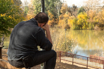 Hombre sentado en el borde de un banco, con expresión pensativa, frente a un río en invierno,...