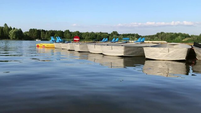 Empty boats and catamarans on the river dock sway on the waves - slow mo shot. Pleasure rowboats, lifeboats and catamarans are moored at the pier on a summer day. Family vacation and tranquility.