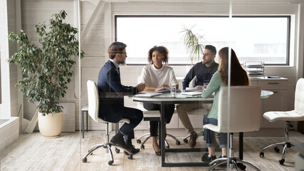 Diverse international businesspeople sit at desk in office boardroom talk discuss ideas together....