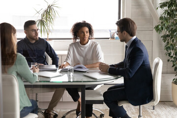 Diverse young businesspeople sit gather in boardroom talk brainstorm at team meeting in office together. Multiracial colleagues coworkers discuss business ideas at briefing. Collaboration concept.