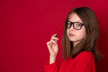 pretty young school girl with glasses and pencil is standing in front of red background