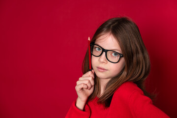 pretty young school girl with glasses and pencil is standing in front of red background