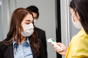 Asian woman watching scanning result from infrared thermometer in colleague hand. checking temperature measurement before enter room at entrance door. Coronavirus prevention, new normal working