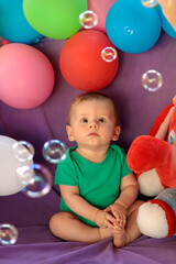 A little kid is sitting in a fun festive atmosphere with balloons and watching soap bubbles.