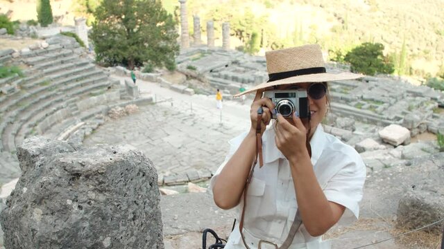 Young woman taking pictures at ruins of temple of Apollo in Delphi, archaeological site by mount Parnassus, Greece, Europe. Vintage camera, large hat, fashion white dress.