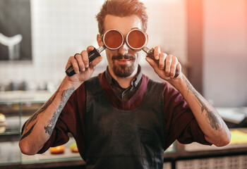 Hipster barista covering eyes with portafilters