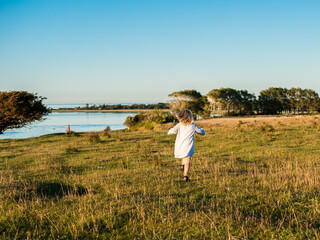 Blond girl 4 years old in white summer dress running and dancing on a field in sunlight and blue sky above