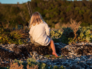 Blond girl in white summer dress sitting with her face turned away on a rock on a beach at sunset while playing with pebbles