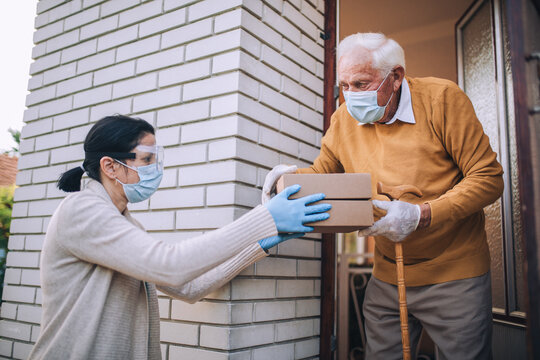 Young Female Volunteer In Mask Gives An Elderly Man Boxes With Food Near His House. Quarantined, Isolated. Coronavirus Covid-19. Donation