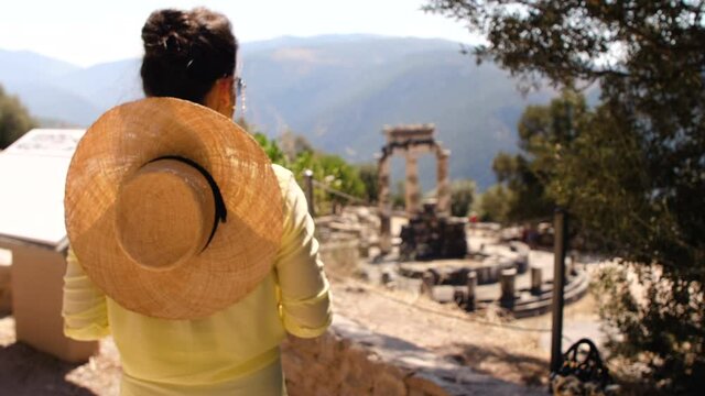 Young woman tourist visiting temple of Athena Pronaia in archaeological site of Delphi, Voiotia, Greece. Fashion elegant yellow dress, large hat.