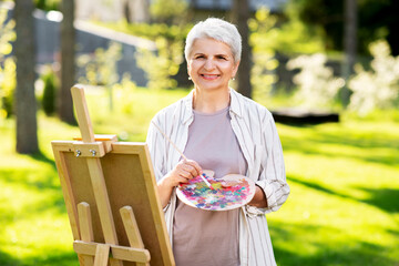 creativity, art and hobby concept - senior woman with easel and color palette painting outdoors