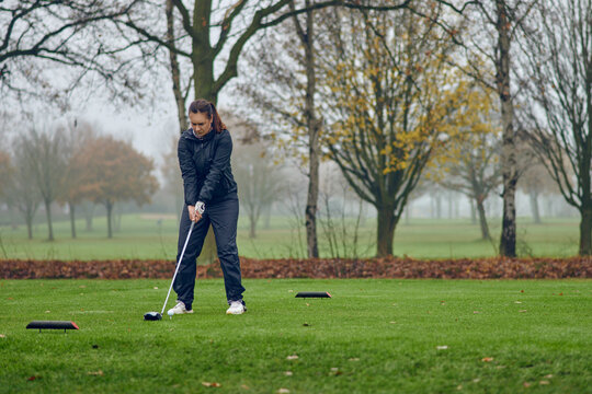 Middle-aged woman playing golf on a cold misty winter day on a parkland course teeing off lining up her shot