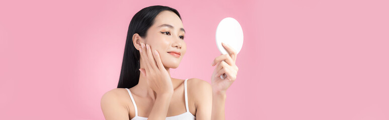 Attractive young Asian woman holding mirror and feeling happy about her skin. Isolated on pink background