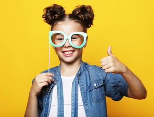 party, childhood and people concept: little girl with a paper accessories over yellow background.