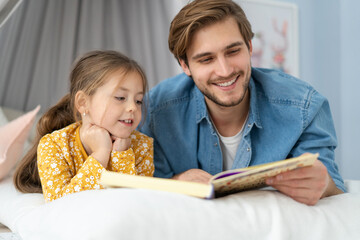 Father reading a book to his daughter while lying on the floor in bedroom.