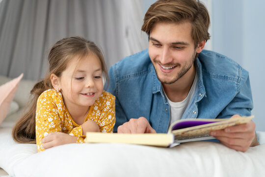 Father reading a book to his daughter while lying on the floor in bedroom.