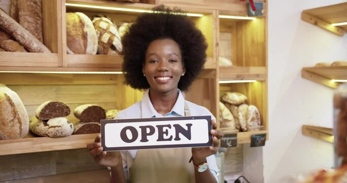Close up portrait of cheerful young African American pretty female seller in apron holding Open sign in hands standing in small bakehouse in good mood on reopening, looking at camera and smiling