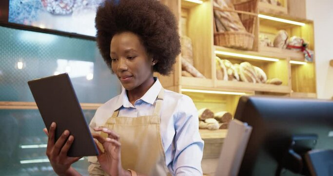 Close up portrait of joyful young African American beautiful female seller worker standing in small bakehouse in good mood typing and scrolling on tablet at workplace. Bakery worker concept