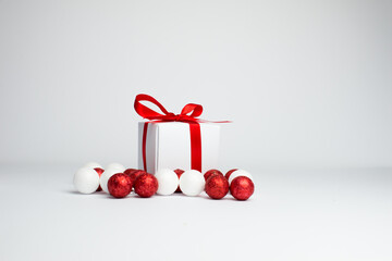 
Festive background. White gift with a red ribbon with white and red christmas balls on a white background. Gift box isolated on white background. Spece for text. Background for Christmas.