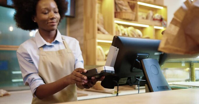 Close up of African American pretty female seller standing in bakery shop at counter typing on computer selling food. Customer hand paying with credit card on device buying baking in bakehouse
