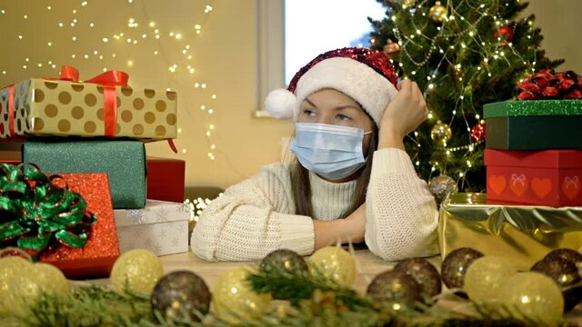 Sad woman in a Santa hat and a protective mask sits alone against the background of a Christmas tree and boxes with gifts. Christmas during the COVID-19 pandemic.