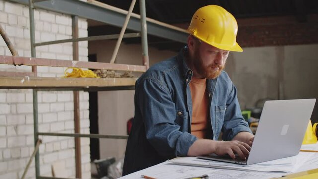 Portrait shot of cheerful male architect in hardhat typing on laptop and desk, then looking at camera and smiling while working at construction site