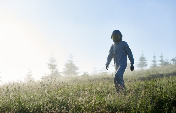 Cosmonaut in space suit walking down the meadow with lush grass. Male astronaut in helmet strolling down grass field with light blue sky on background. Concept of astronautics, exploration and nature.