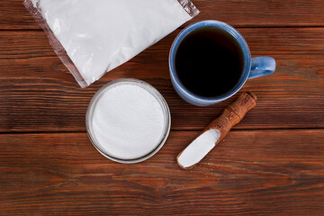 Fructose in a transparent pack with ceramic cup of tea, glass jar and wooden scoop of sorbitol on a wooden background. Different sweeteners concept. Top view.