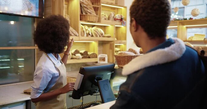 Rear of African American man client buying bread and baking in bakehouse. Beautiful happy young female worker seller selling fresh bread in bakery shop. Small business concept