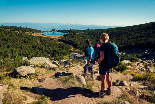 Bulgarian man and Polish woman tourists with backpacks in Pirin national park looking at Bezbog hut and Bezbozhko Lake in sunny summer day
