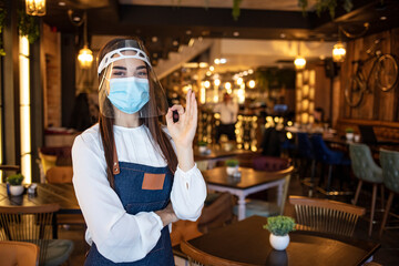 Fototapeta na wymiar Portrait attractive waitress wear face mask and face shield showing OK sign with indoor restaurant background. New normal restaurant concept. Beautiful waitress working at a cafe