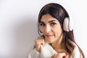 Listening to music in headphones. Portrait of beautiful brunette woman in comfortable soft longsleeve isolated on white studio background. Home comfort, emotions, winter mood concept. Copyspace.